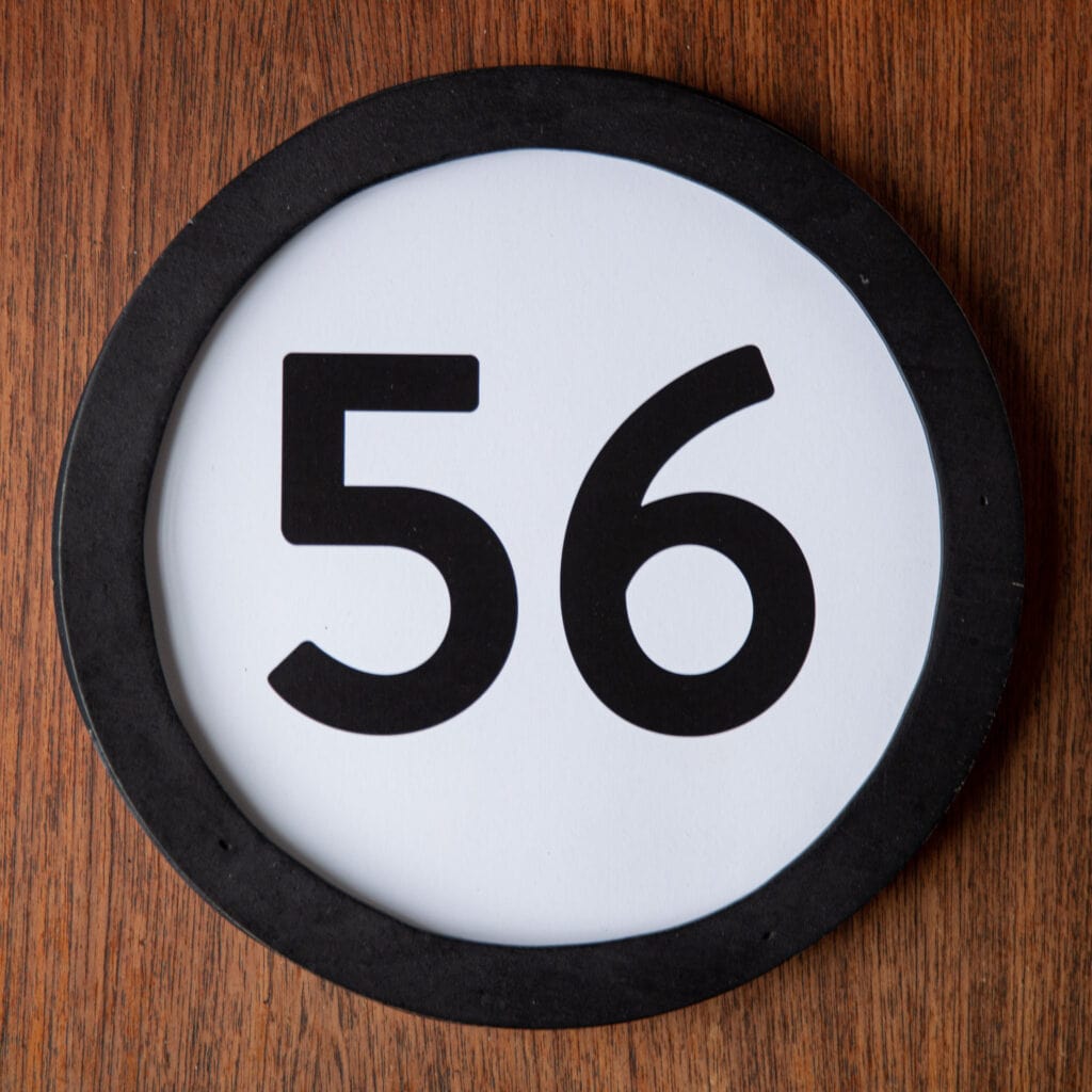 A circle shaped token fills the square image. The token has a 1 inch black circular frame around it. In the center is a black number on a white background. The token lies on a teak wood table. This digital image is part of the 1 to Infinity portrait photography series by Danny Goldfield.