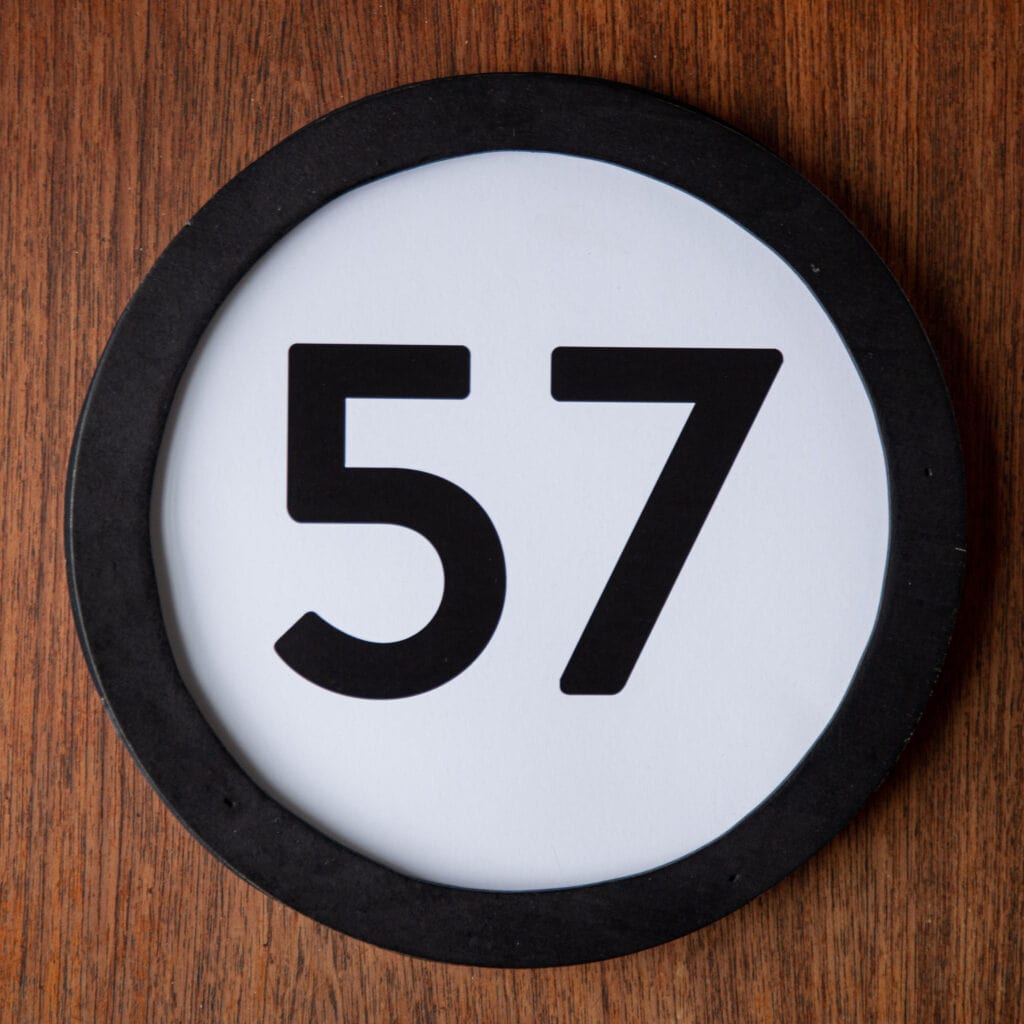 A circle shaped token fills the square image. The token has a 1 inch black circular frame around it. In the center is a black number on a white background. The token lies on a teak wood table. This digital image is part of the 1 to Infinity portrait photography series by Danny Goldfield.