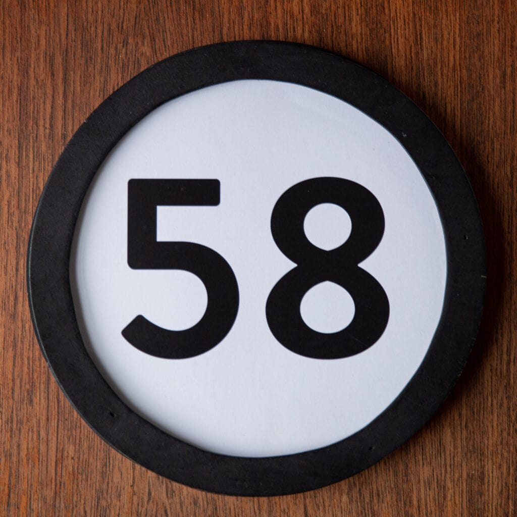 A circle shaped token fills the square image. The token has a 1 inch black circular frame around it. In the center is a black number on a white background. The token lies on a teak wood table. This digital image is part of the 1 to Infinity portrait photography series by Danny Goldfield.