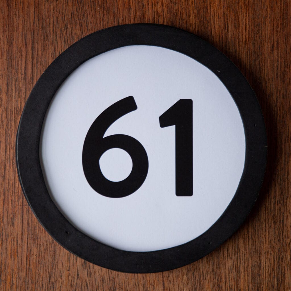 A circle shaped token fills the square image. The token has a 1 inch black circular frame around it. In the center is a black number on a white background. The token lies on a teak wood table. This digital image is part of the 1 to Infinity portrait photography series by Danny Goldfield.