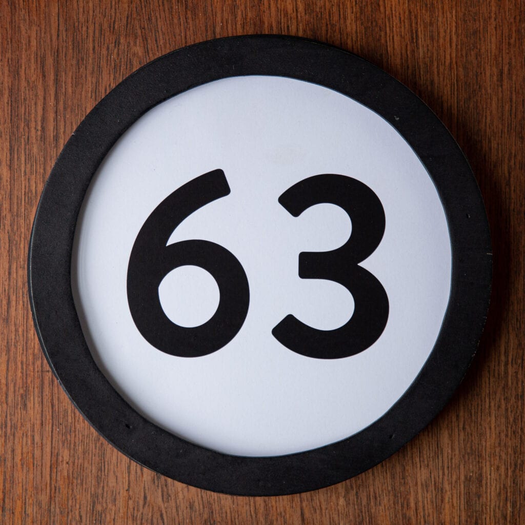 A circle shaped token fills the square image. The token has a 1 inch black circular frame around it. In the center is a black number on a white background. The token lies on a teak wood table. This digital image is part of the 1 to Infinity portrait photography series by Danny Goldfield.