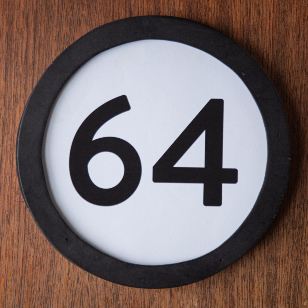 A circle shaped token fills the square image. The token has a 1 inch black circular frame around it. In the center is a black number on a white background. The token lies on a teak wood table. This digital image is part of the 1 to Infinity portrait photography series by Danny Goldfield.