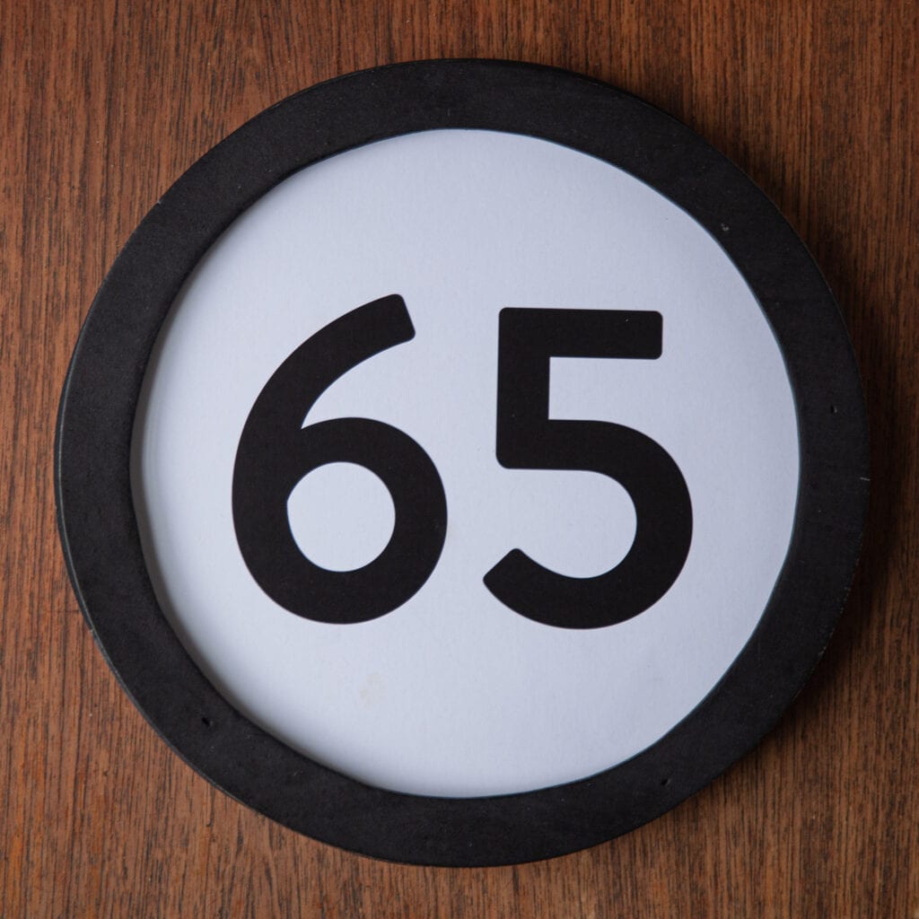 A circle shaped token fills the square image. The token has a 1 inch black circular frame around it. In the center is a black number on a white background. The token lies on a teak wood table. This digital image is part of the 1 to Infinity portrait photography series by Danny Goldfield.