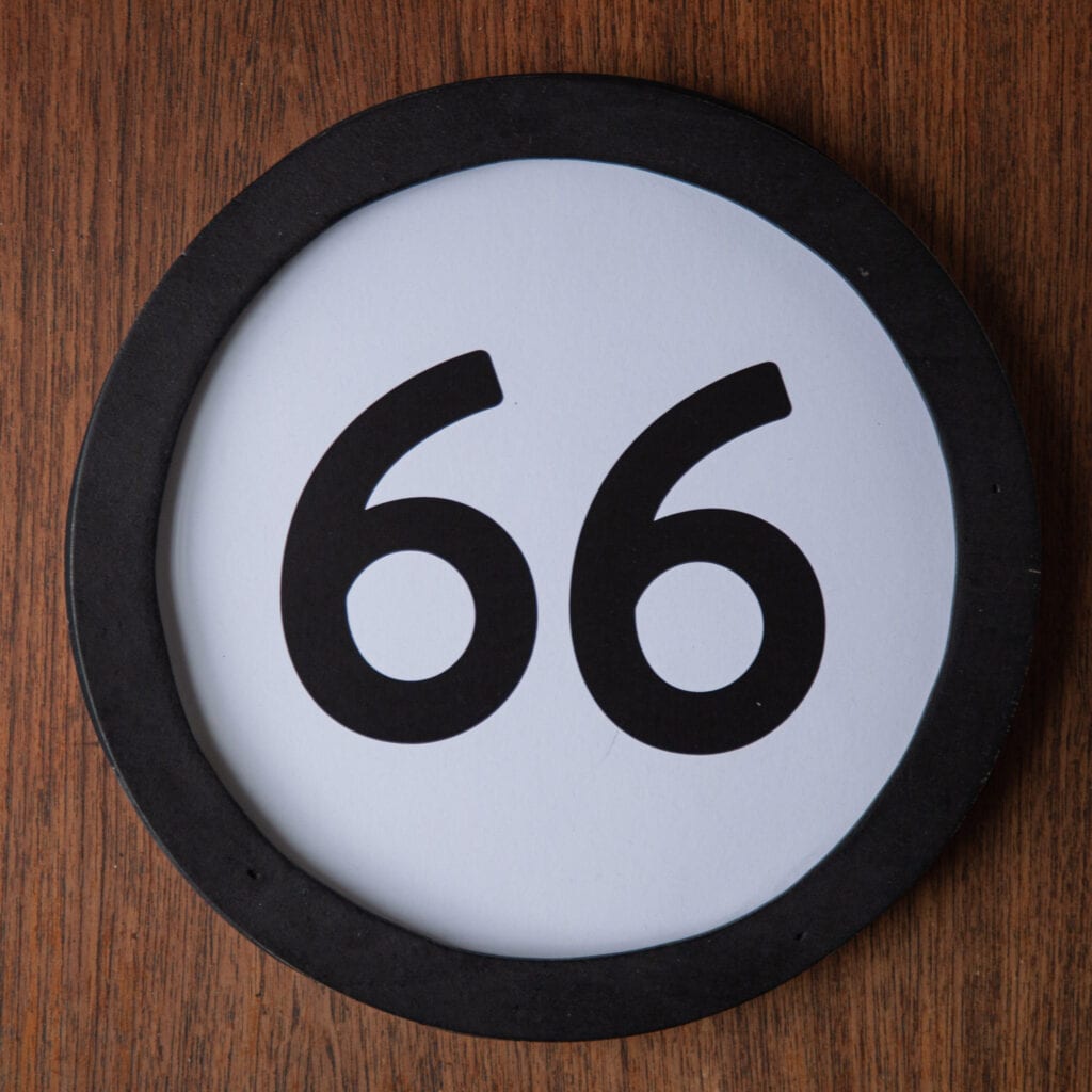 A circle shaped token fills the square image. The token has a 1 inch black circular frame around it. In the center is a black number on a white background. The token lies on a teak wood table. This digital image is part of the 1 to Infinity portrait photography series by Danny Goldfield.