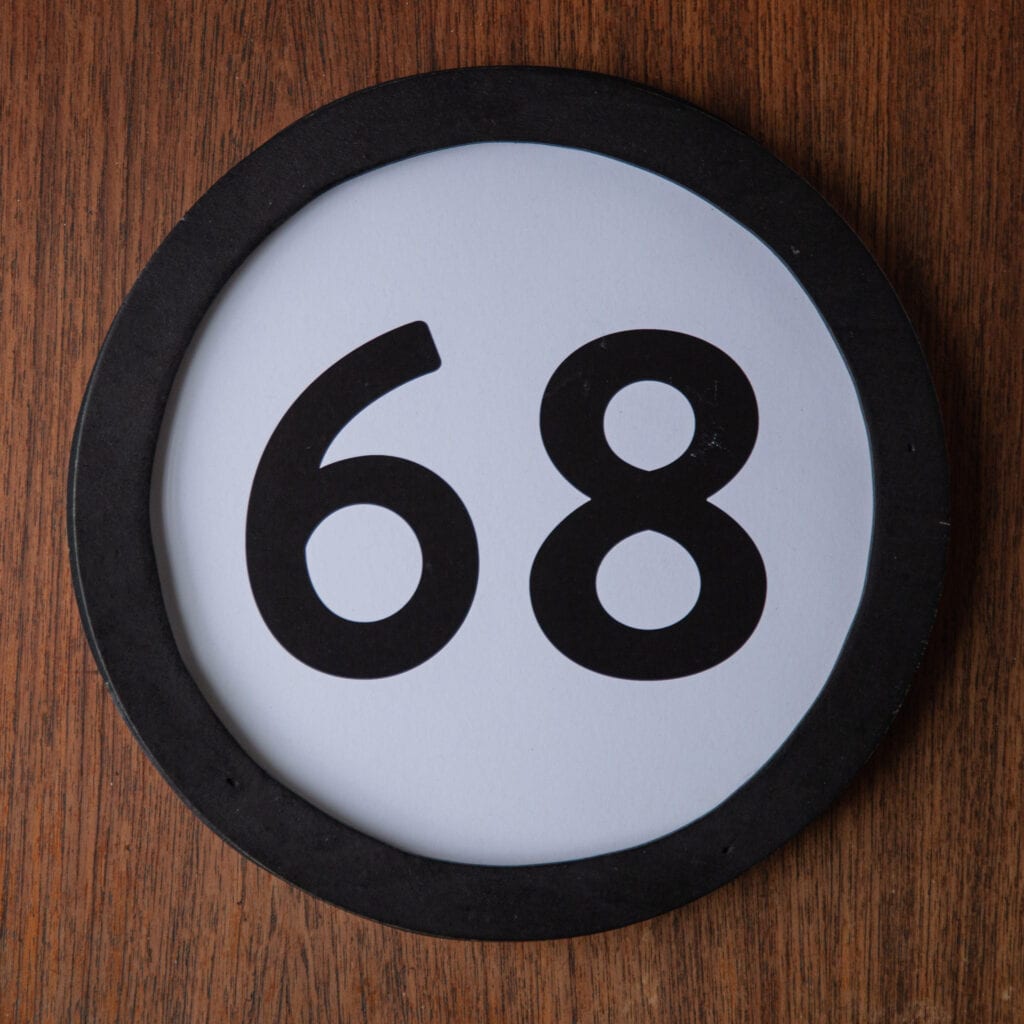 A circle shaped token fills the square image. The token has a 1 inch black circular frame around it. In the center is a black number on a white background. The token lies on a teak wood table. This digital image is part of the 1 to Infinity portrait photography series by Danny Goldfield.