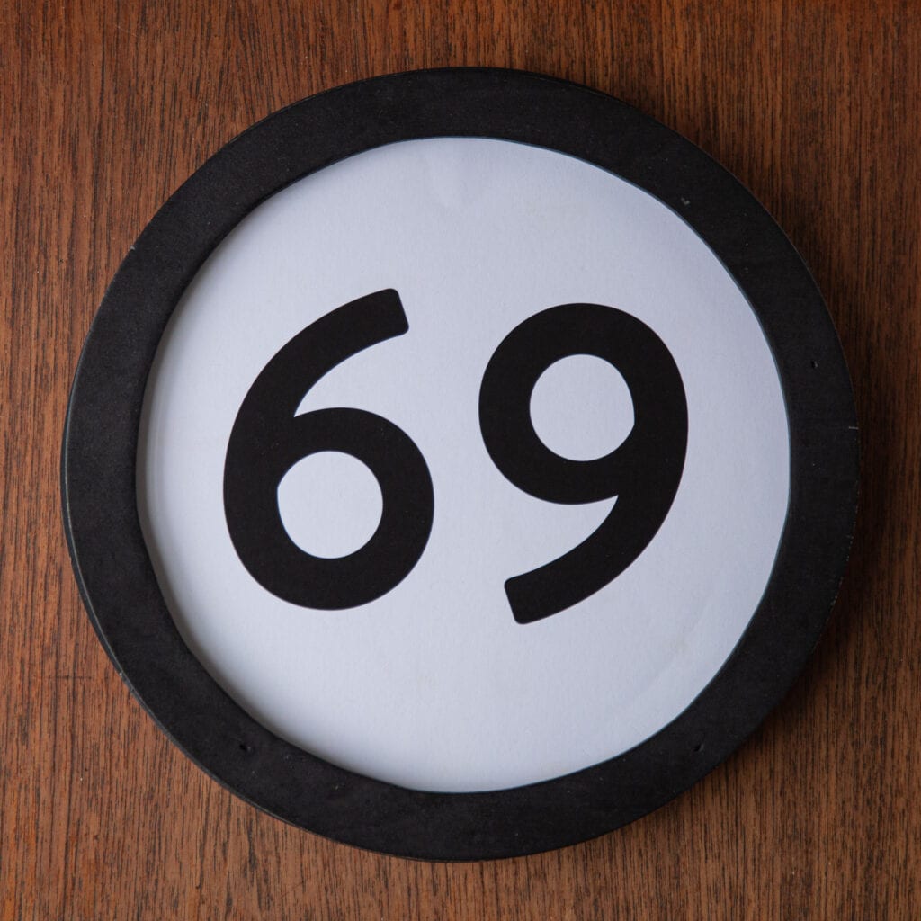 A circle shaped token fills the square image. The token has a 1 inch black circular frame around it. In the center is a black number on a white background. The token lies on a teak wood table. This digital image is part of the 1 to Infinity portrait photography series by Danny Goldfield.