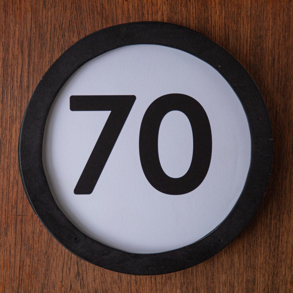 A circle shaped token fills the square image. The token has a 1 inch black circular frame around it. In the center is a black number on a white background. The token lies on a teak wood table. This digital image is part of the 1 to Infinity portrait photography series by Danny Goldfield.