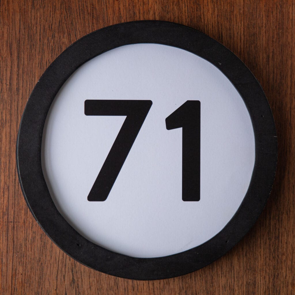 A circle shaped token fills the square image. The token has a 1 inch black circular frame around it. In the center is a black number on a white background. The token lies on a teak wood table. This digital image is part of the 1 to Infinity portrait photography series by Danny Goldfield.