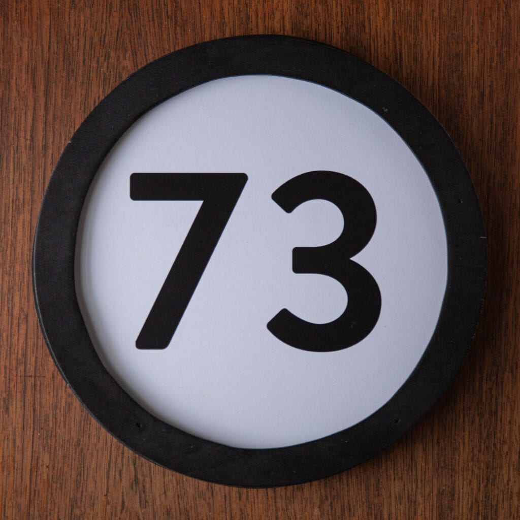 A circle shaped token fills the square image. The token has a 1 inch black circular frame around it. In the center is a black number on a white background. The token lies on a teak wood table. This digital image is part of the 1 to Infinity portrait photography series by Danny Goldfield.