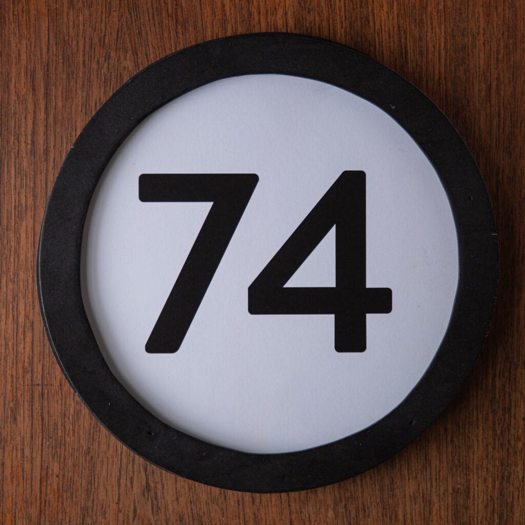 A circle shaped token fills the square image. The token has a 1 inch black circular frame around it. In the center is a black number on a white background. The token lies on a teak wood table. This digital image is part of the 1 to Infinity portrait photography series by Danny Goldfield.