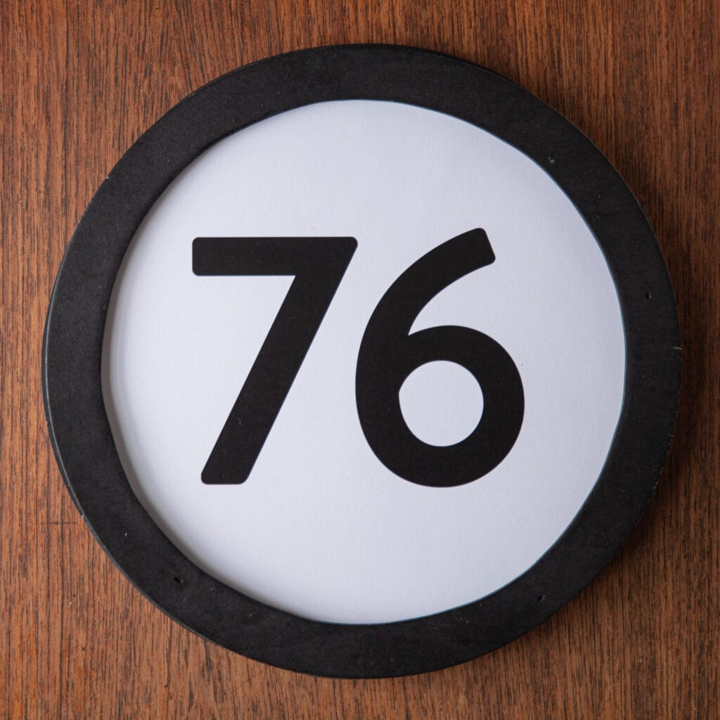 A circle shaped token fills the square image. The token has a 1 inch black circular frame around it. In the center is a black number on a white background. The token lies on a teak wood table. This digital image is part of the 1 to Infinity portrait photography series by Danny Goldfield.