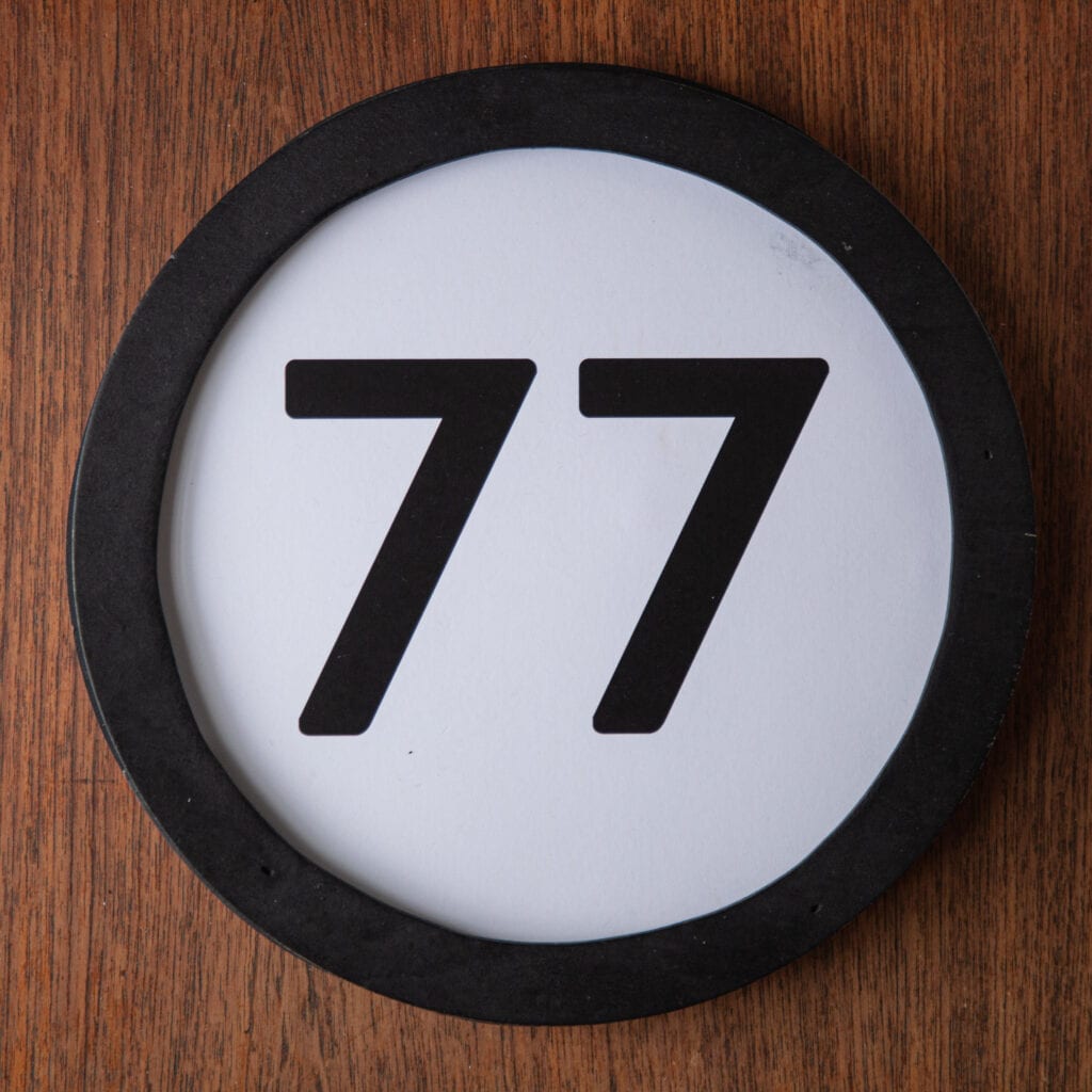 A circle shaped token fills the square image. The token has a 1 inch black circular frame around it. In the center is a black number on a white background. The token lies on a teak wood table. This digital image is part of the 1 to Infinity portrait photography series by Danny Goldfield.