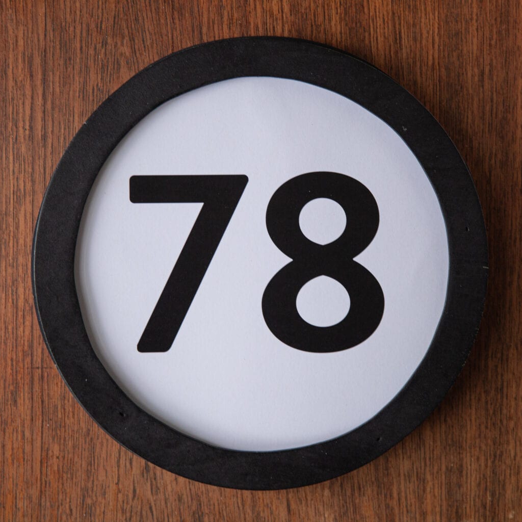A circle shaped token fills the square image. The token has a 1 inch black circular frame around it. In the center is a black number on a white background. The token lies on a teak wood table. This digital image is part of the 1 to Infinity portrait photography series by Danny Goldfield.