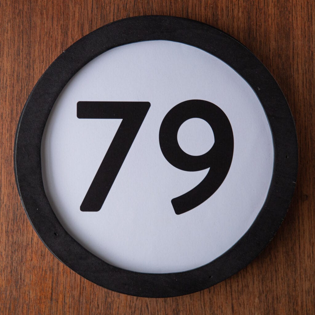 A circle shaped token fills the square image. The token has a 1 inch black circular frame around it. In the center is a black number on a white background. The token lies on a teak wood table. This digital image is part of the 1 to Infinity portrait photography series by Danny Goldfield.