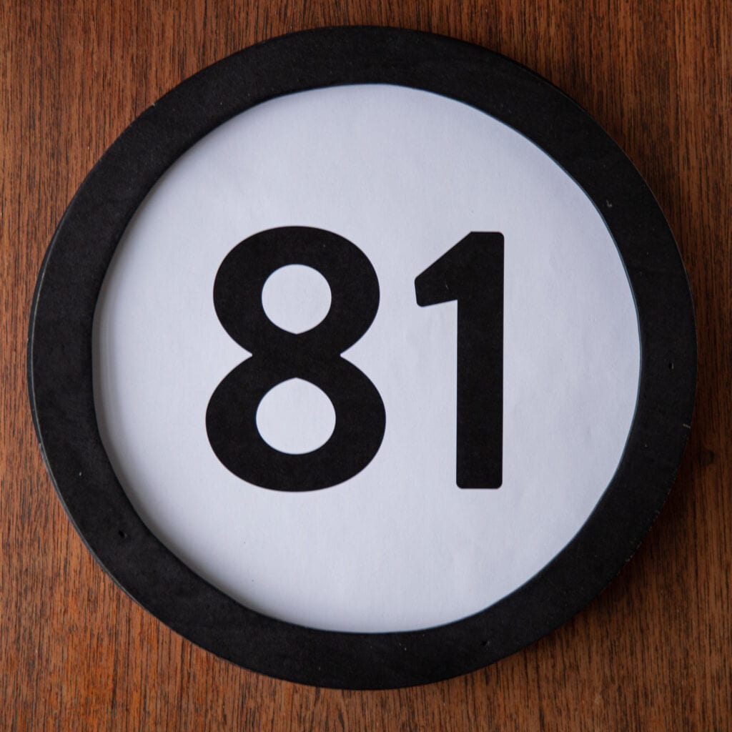 A circle shaped token fills the square image. The token has a 1 inch black circular frame around it. In the center is a black number on a white background. The token lies on a teak wood table. This digital image is part of the 1 to Infinity portrait photography series by Danny Goldfield.