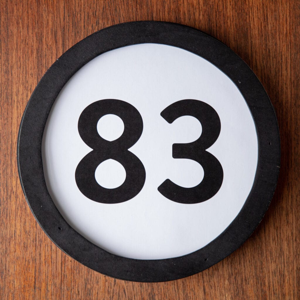 A circle shaped token fills the square image. The token has a 1 inch black circular frame around it. In the center is a black number on a white background. The token lies on a teak wood table. This digital image is part of the 1 to Infinity portrait photography series by Danny Goldfield.