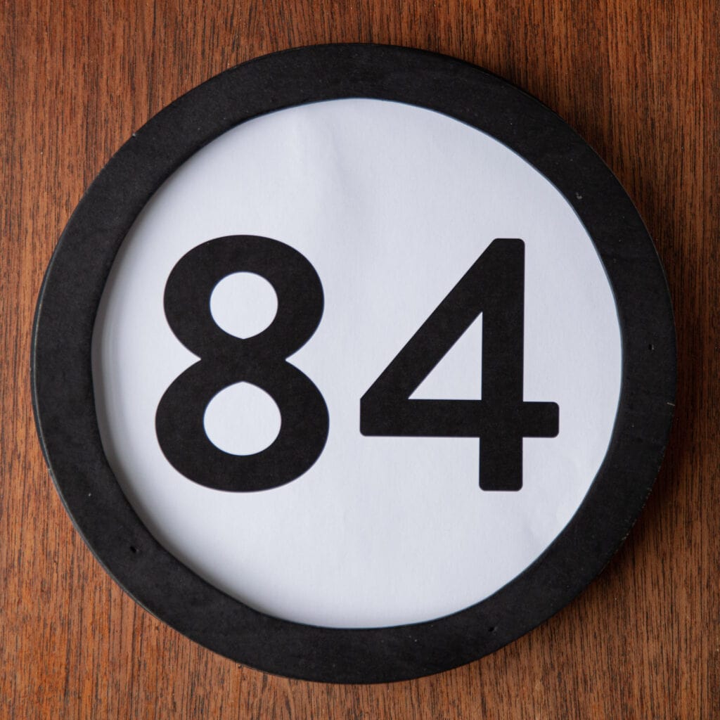A circle shaped token fills the square image. The token has a 1 inch black circular frame around it. In the center is a black number on a white background. The token lies on a teak wood table. This digital image is part of the 1 to Infinity portrait photography series by Danny Goldfield.