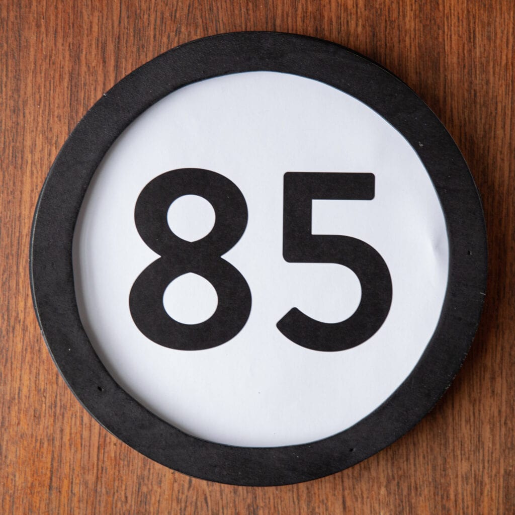 A circle shaped token fills the square image. The token has a 1 inch black circular frame around it. In the center is a black number on a white background. The token lies on a teak wood table. This digital image is part of the 1 to Infinity portrait photography series by Danny Goldfield.