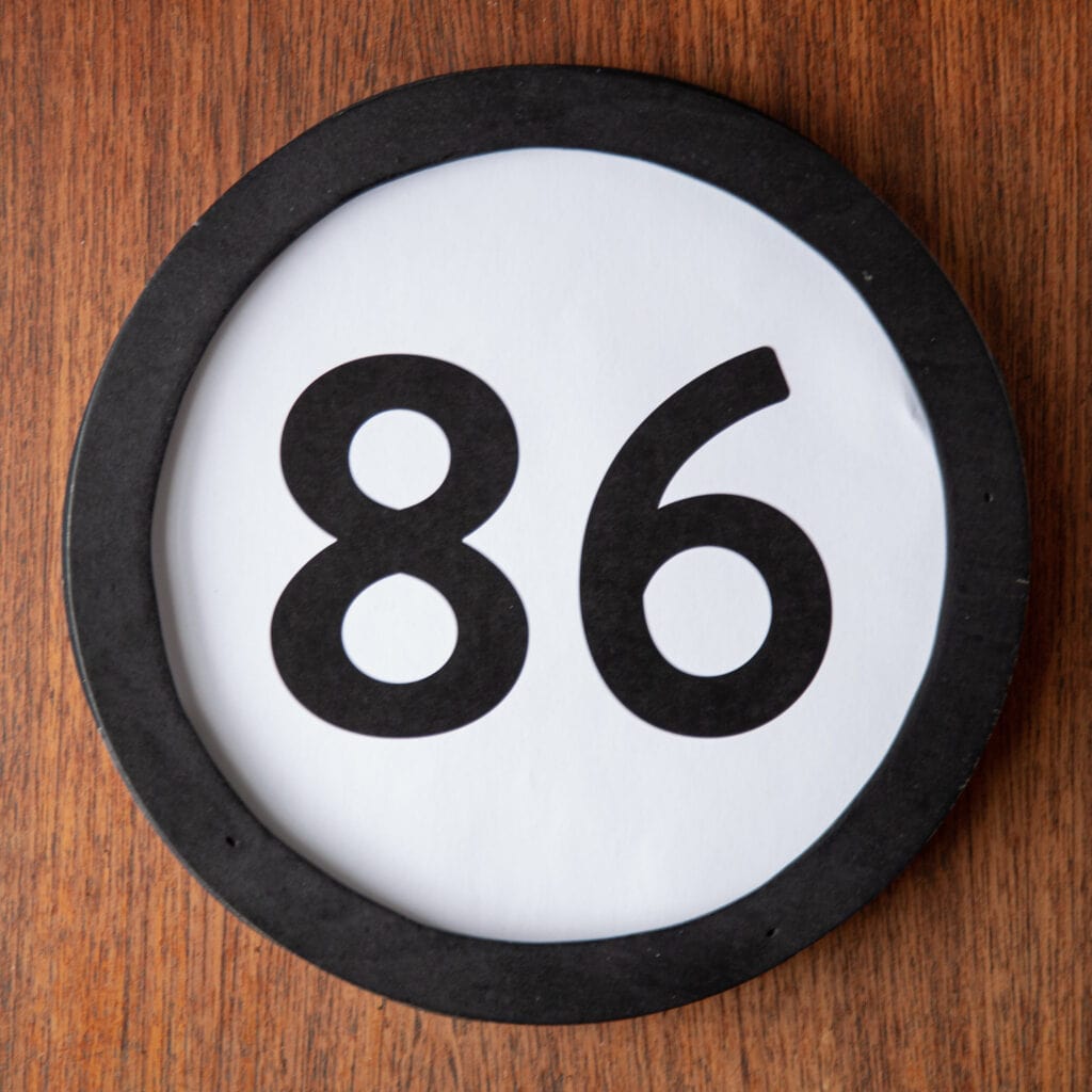 A circle shaped token fills the square image. The token has a 1 inch black circular frame around it. In the center is a black number on a white background. The token lies on a teak wood table. This digital image is part of the 1 to Infinity portrait photography series by Danny Goldfield.