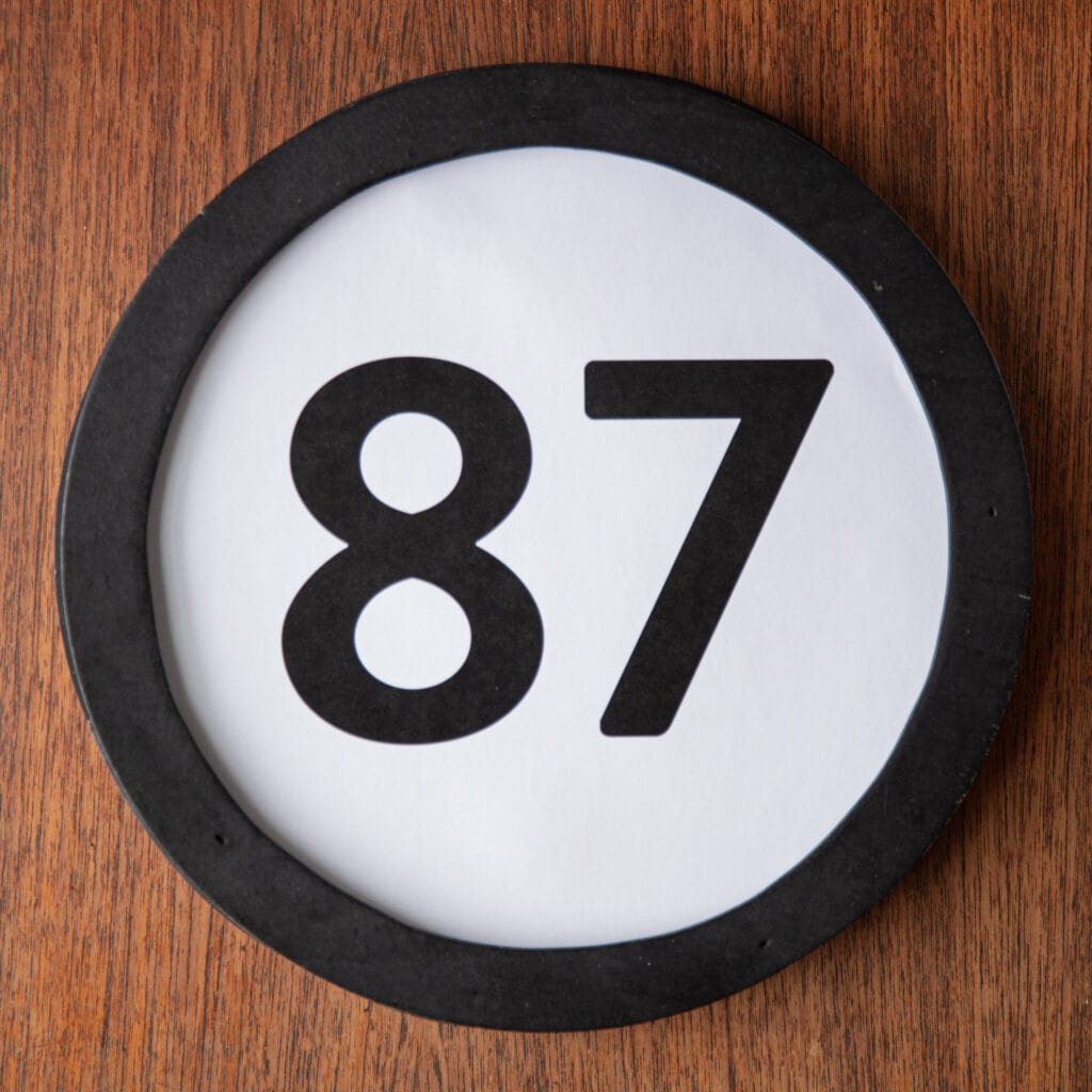 A circle shaped token fills the square image. The token has a 1 inch black circular frame around it. In the center is a black number on a white background. The token lies on a teak wood table. This digital image is part of the 1 to Infinity portrait photography series by Danny Goldfield.