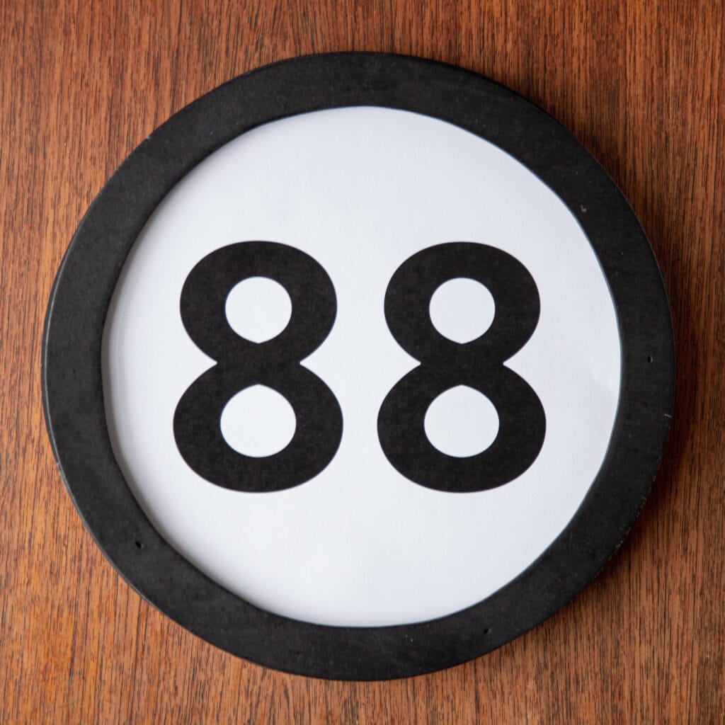 A circle shaped token fills the square image. The token has a 1 inch black circular frame around it. In the center is a black number on a white background. The token lies on a teak wood table. This digital image is part of the 1 to Infinity portrait photography series by Danny Goldfield.