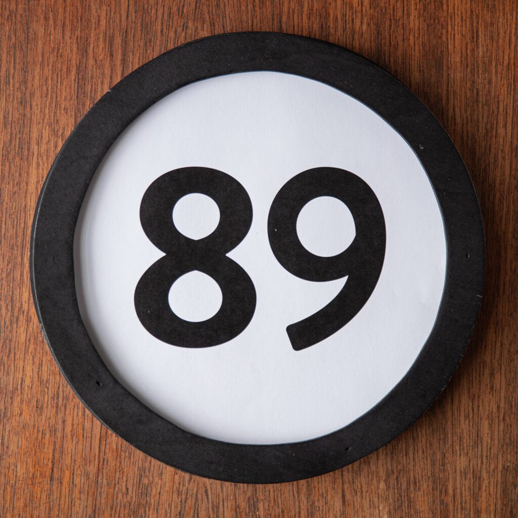 A circle shaped token fills the square image. The token has a 1 inch black circular frame around it. In the center is a black number on a white background. The token lies on a teak wood table. This digital image is part of the 1 to Infinity portrait photography series by Danny Goldfield.