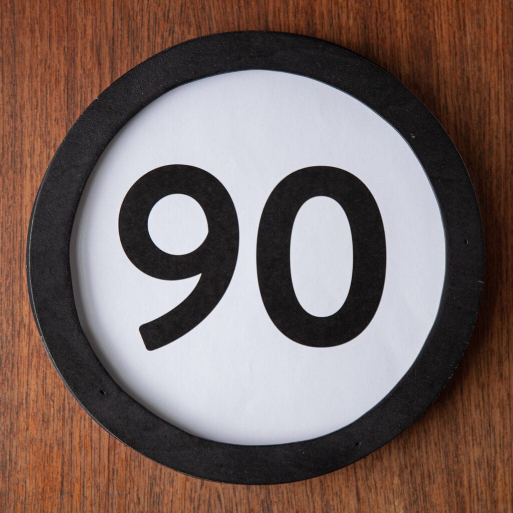 A circle shaped token fills the square image. The token has a 1 inch black circular frame around it. In the center is a black number on a white background. The token lies on a teak wood table. This digital image is part of the 1 to Infinity portrait photography series by Danny Goldfield.