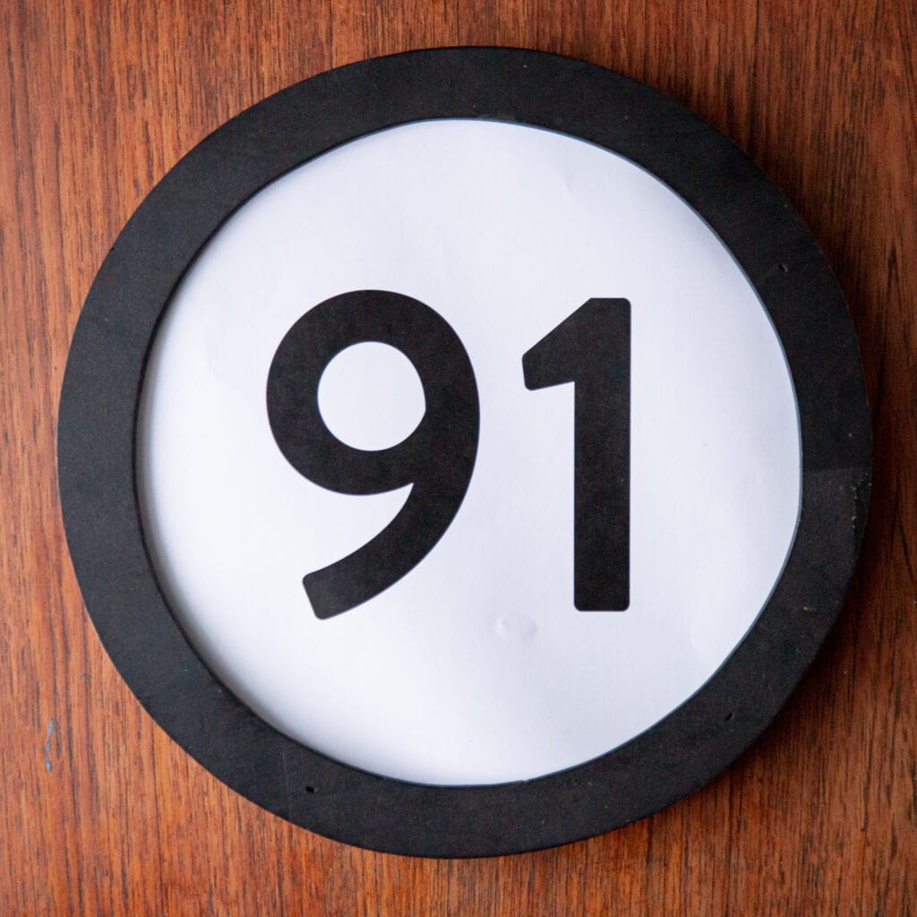 A circle shaped token fills the square image. The token has a 1 inch black circular frame around it. In the center is a black number on a white background. The token lies on a teak wood table. This digital image is part of the 1 to Infinity portrait photography series by Danny Goldfield.