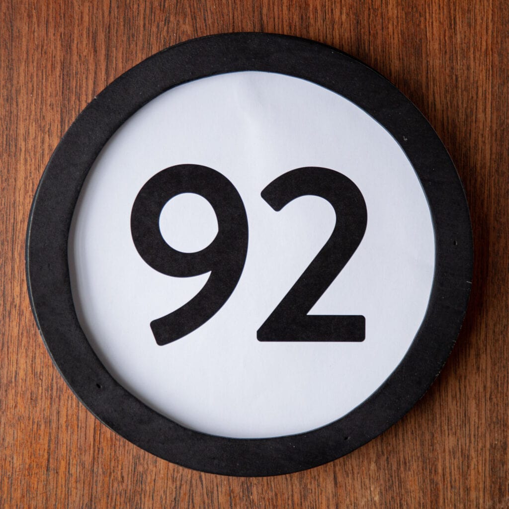 A circle shaped token fills the square image. The token has a 1 inch black circular frame around it. In the center is a black number on a white background. The token lies on a teak wood table. This digital image is part of the 1 to Infinity portrait photography series by Danny Goldfield.