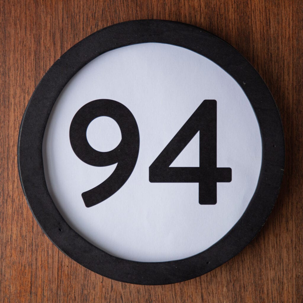 A circle shaped token fills the square image. The token has a 1 inch black circular frame around it. In the center is a black number on a white background. The token lies on a teak wood table. This digital image is part of the 1 to Infinity portrait photography series by Danny Goldfield.