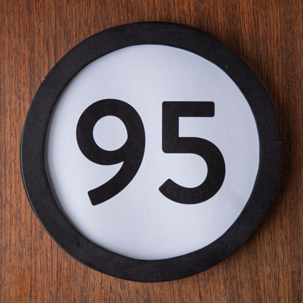 A circle shaped token fills the square image. The token has a 1 inch black circular frame around it. In the center is a black number on a white background. The token lies on a teak wood table. This digital image is part of the 1 to Infinity portrait photography series by Danny Goldfield.