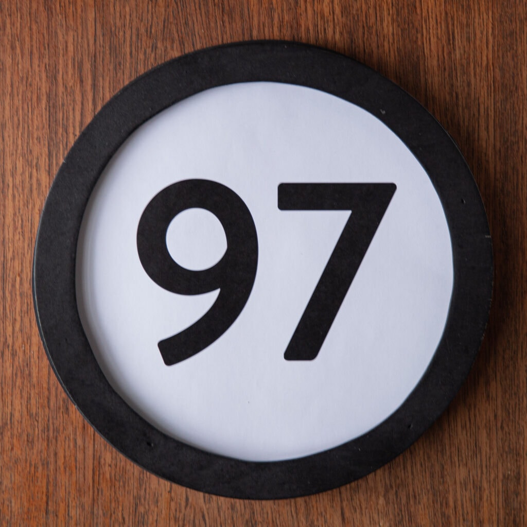 A circle shaped token fills the square image. The token has a 1 inch black circular frame around it. In the center is a black number on a white background. The token lies on a teak wood table. This digital image is part of the 1 to Infinity portrait photography series by Danny Goldfield.
