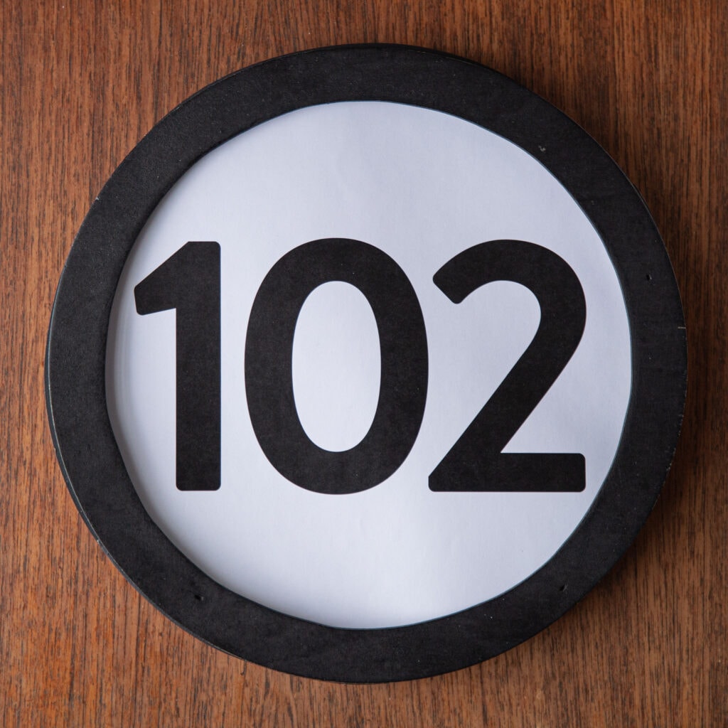 A circle shaped token fills the square image. The token has a 1 inch black circular frame around it. In the center is a black number on a white background. The token lies on a teak wood table. This digital image is part of the 1 to Infinity portrait photography series by Danny Goldfield.
