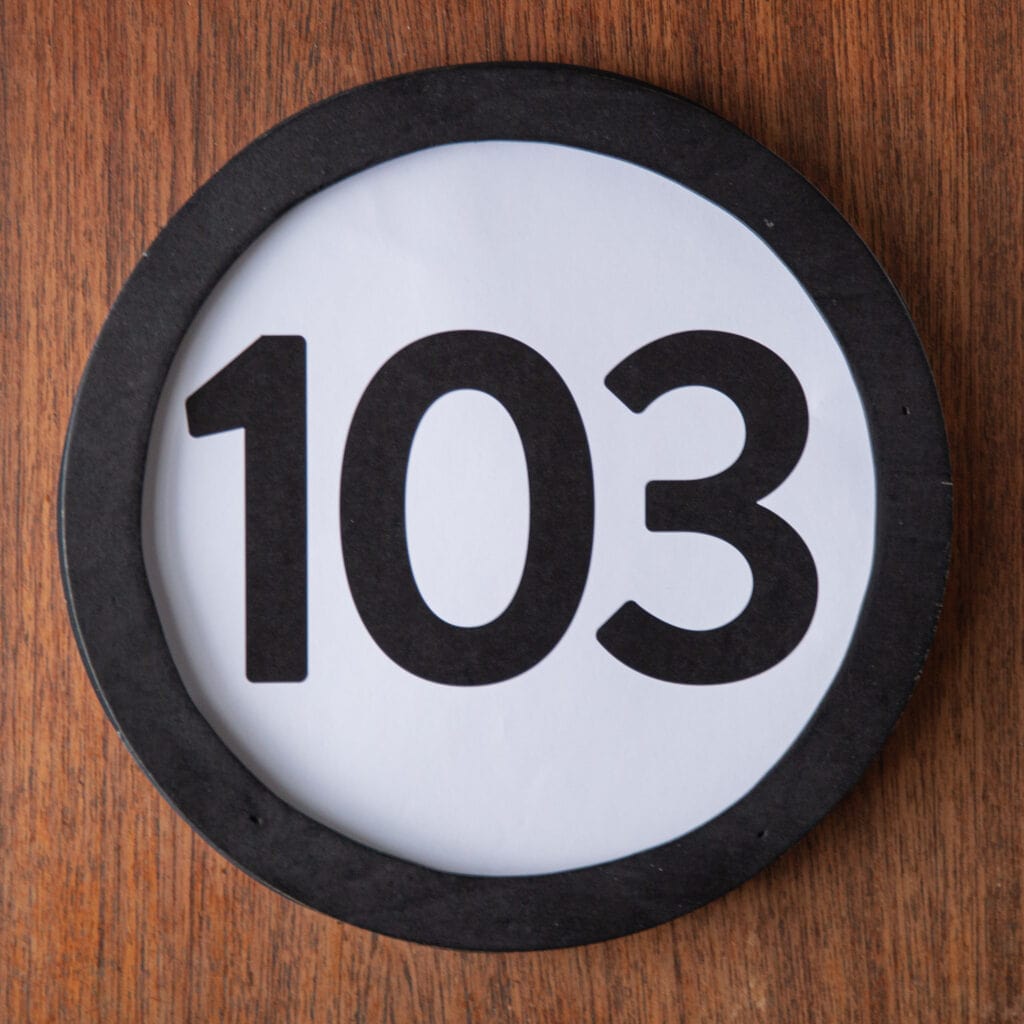 A circle shaped token fills the square image. The token has a 1 inch black circular frame around it. In the center is a black number on a white background. The token lies on a teak wood table. This digital image is part of the 1 to Infinity portrait photography series by Danny Goldfield.
