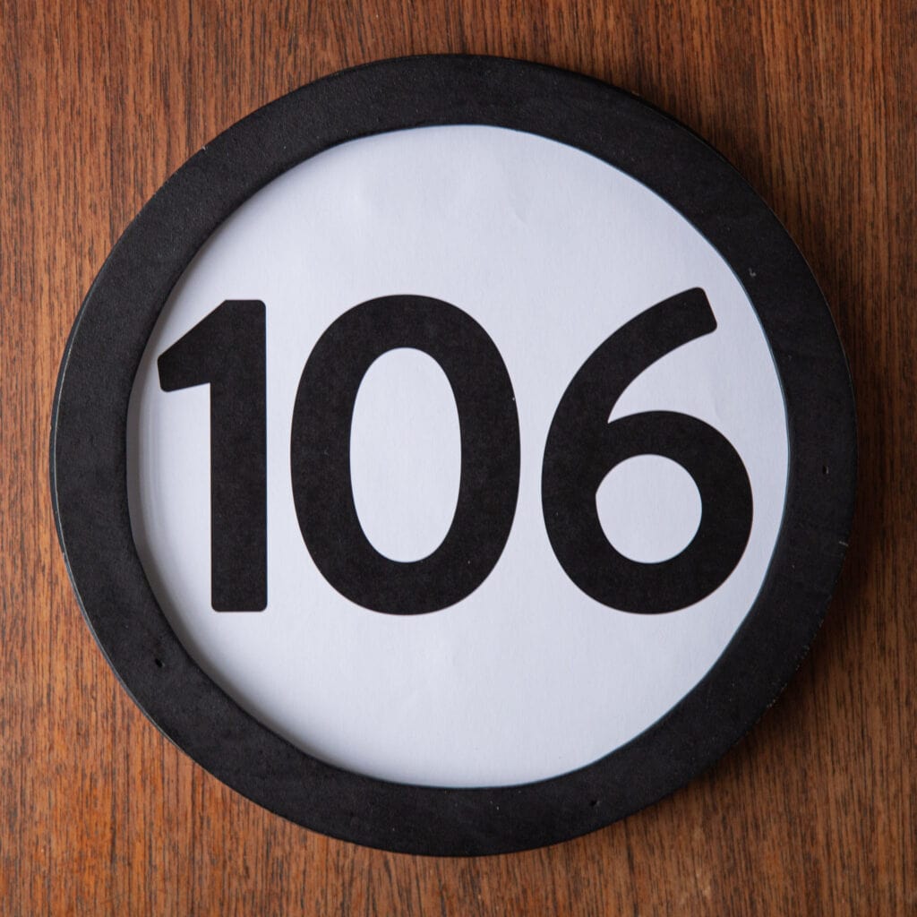 A circle shaped token fills the square image. The token has a 1 inch black circular frame around it. In the center is a black number on a white background. The token lies on a teak wood table. This digital image is part of the 1 to Infinity portrait photography series by Danny Goldfield.