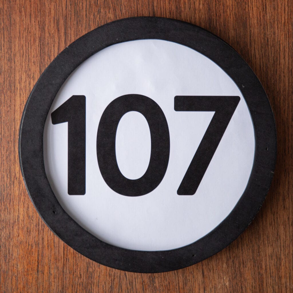 A circle shaped token fills the square image. The token has a 1 inch black circular frame around it. In the center is a black number on a white background. The token lies on a teak wood table. This digital image is part of the 1 to Infinity portrait photography series by Danny Goldfield.